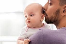 Baby wearing a white onesie with pink trim being held by an adult in a purple shirt.